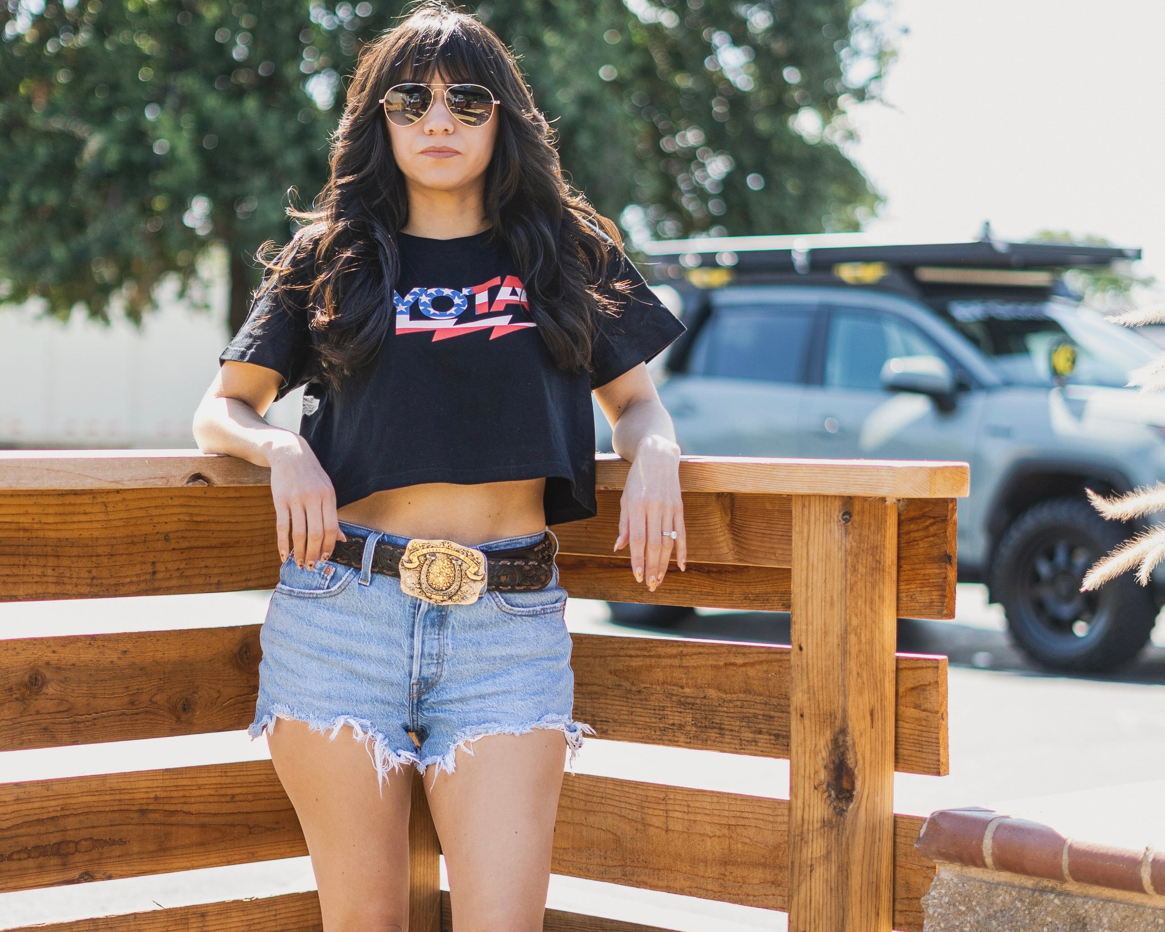 Woman wearing a black crop top with red text, denim shorts, and brown cowboy boots standing on a wooden platform.